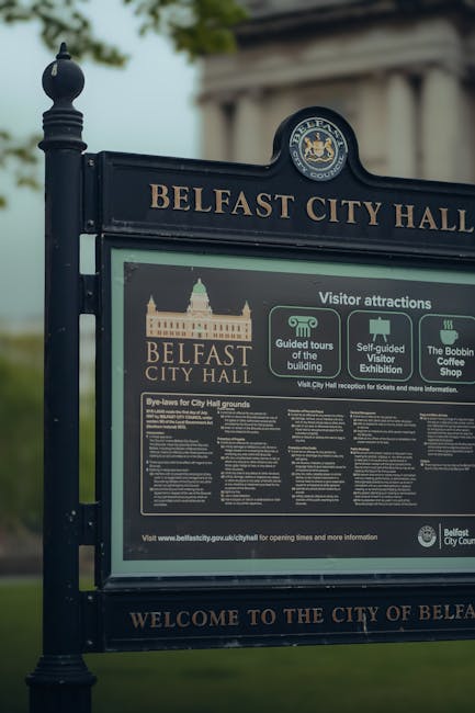 A black, decorative information board with the heading 'Belfast City Hall' in gold lettering, situated outdoors on a paved or grassy area, with a background of blurred green trees and a cloudy sky. The board features the Belfast City Hall emblem at the top, along with sections providing visitor information, including guided tour details, self-guided visitor exhibition, and the Bobbin Coffee Shop. Below these sections, there is a larger, densely printed block of text titled 'Bye-laws for City Hall grounds' with detailed rules. The surroundings suggest a park or outdoor public space, and the sign appears to be part of a public or civic area, potentially managed with independent waste collection or local authority services. The overall scene is calm with natural lighting, emphasizing the informational purpose of the display, while subtly relating to the importance of maintaining clean, well-managed public environments, possibly through private rubbish removal or site clearance services offered by Rubbish Collection Kennington.