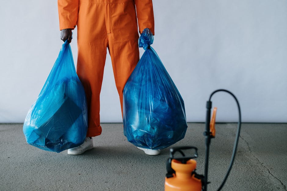 A person wearing an orange work jumpsuit stands on a grey concrete floor, holding two large blue plastic rubbish bags filled with waste, one in each hand. The bags are slightly translucent, revealing contents such as crumpled paper and lightweight debris, and are tied at the top. The individual's white shoes are visible at the bottom of the image. In the foreground, a black and orange handheld sprayer or vacuum device is partially visible, with a flexible black hose extending towards the top right of the frame. The background is a plain, light grey wall, creating a neutral setting typical of a waste collection or disposal area. The scene emphasizes the manual handling of waste, aligning with private rubbish removal services that provide on-site collection of rubbish from residential or commercial properties, such as flats or apartments, in an urban environment.