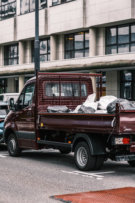 A small burgundy flatbed pickup truck parked on an urban street near a modern office building with large glass windows and a smooth, light-colored concrete facade. The truck's cargo bed is filled with various types of waste, including black and white plastic bags, cardboard boxes, and other loose rubbish materials, some of which are slightly disorganized and protruding from the bed. The truck features a protective metal grid at the rear for securing the load, and its body shows signs of regular use, with visible minor wear and a clean finish. The scene is illuminated by daylight, with shadows cast on the pavement, and a streetlight pole is visible behind the vehicle. This image, associated with Rubbish Collection Kennington, exemplifies private waste removal services specializing in on-site clearance of rubbish from commercial or residential buildings, supporting alternative waste handling methods outside municipal collection systems.