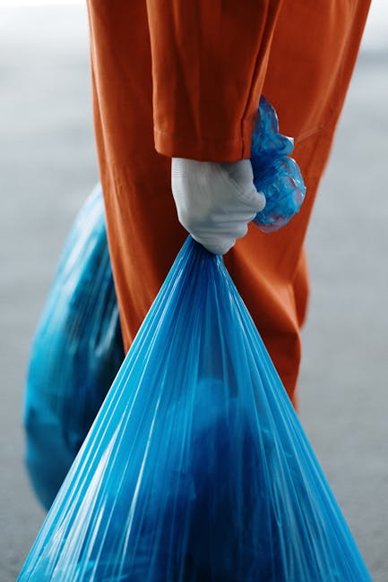 A person wearing an orange work jumpsuit stands on a grey concrete floor, holding two large blue plastic rubbish bags filled with waste, one in each hand. The bags are slightly translucent, revealing contents such as crumpled paper and lightweight debris, and are tied at the top. The individual's white shoes are visible at the bottom of the image. In the foreground, a black and orange handheld sprayer or vacuum device is partially visible, with a flexible black hose extending towards the top right of the frame. The background is a plain, light grey wall, creating a neutral setting typical of a waste collection or disposal area. The scene emphasizes the manual handling of waste, aligning with private rubbish removal services that provide on-site collection of rubbish from residential or commercial properties, such as flats or apartments, in an urban environment.