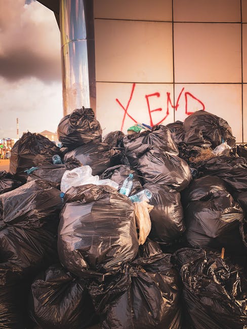 A large pile of black bin bags filled with waste is stacked against a modern, tiled building wall, with some white plastic bottles and other discarded items visible on top or around the bags. The bags are tightly knotted and appear to contain various rubbish, with a few plastic bottles and pieces of paper peeking out from the openings. In the background, part of a cityscape with buildings and a cloudy sky is visible, suggesting an urban environment. The wall behind the waste display features large, beige tiles with visible grout lines, and there is red graffiti text that reads 'XEN' spray-painted on the wall. The scene is lit by natural daylight, casting shadows on the bags, and the overall impression indicates an instance of informal or unscheduled rubbish accumulation, possibly related to private waste handling or on-site clearance carried out by a local waste management service such as Rubbish Collection Kennington, showcasing typical challenges with rubbish removal in public or commercial spaces.