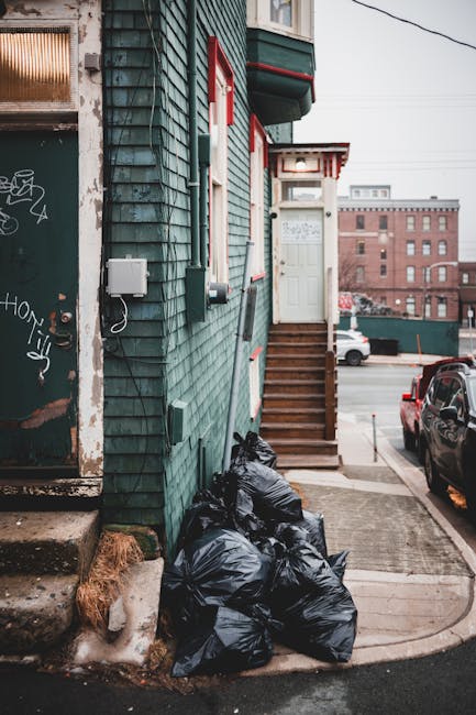 A collection of black rubbish bags, tightly tied and grouped, are stacked at the base of a weathered wooden staircase outside a building with green wooden siding. The building features red and white trim around a small upstairs porch with a white door and stairs. To the left, a graffiti-covered metal door is partially visible, attached to the edge of the building, with a small utility box mounted on the wall nearby. The scene is set on a paved sidewalk adjacent to a street with moving and parked vehicles, including a dark-colored car. In the background, a brick industrial-style building is visible under an overcast sky, and electrical wires run above the scene. The environment suggests an urban area where independent waste collection services, such as those offered by Rubbish Collection Kennington, may handle rubbish and on-site clearance outside residential buildings, especially in situations where waste disposal through local authority services is not utilised.