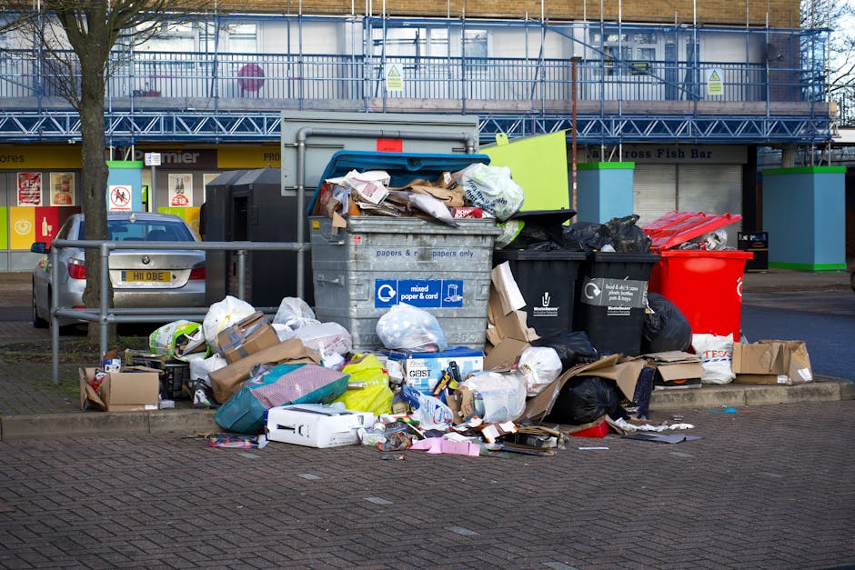A large pile of mixed waste and rubbish bags is accumulated on a raised sidewalk section in front of a commercial area. The collection includes black plastic refuse bags, cardboard boxes, paper, and packaging materials, some of which are torn open or spilling onto the paving. There are also a few white plastic bags and loose debris scattered around the pile. Behind the rubbish, a commercial building with a scaffolding-covered facade and a cantilevered balcony is visible, with signage indicating shops and a fish bar. To the left, a parked silver car with a partially visible yellow license plate faces away from the camera. The scene is outdoors, with a metal railing separating the rubbish from the vehicle and pedestrian areas. The environment appears to be an urban or suburban street scene, and the image reflects the type of waste accumulation that private waste removal services, such as those offered by Rubbish Collection Kennington, might be called upon to clear, especially outside of standard council waste collection zones. The lighting is natural, suggesting daytime, and the overall scene is neutral, highlighting the typical appearance of uncollected or overflowing rubbish on a street corner.