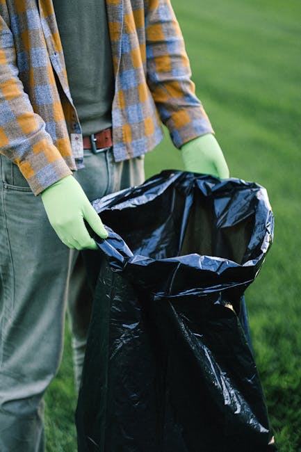 A large pile of mixed waste and rubbish bags is accumulated on a raised sidewalk section in front of a commercial area. The collection includes black plastic refuse bags, cardboard boxes, paper, and packaging materials, some of which are torn open or spilling onto the paving. There are also a few white plastic bags and loose debris scattered around the pile. Behind the rubbish, a commercial building with a scaffolding-covered facade and a cantilevered balcony is visible, with signage indicating shops and a fish bar. To the left, a parked silver car with a partially visible yellow license plate faces away from the camera. The scene is outdoors, with a metal railing separating the rubbish from the vehicle and pedestrian areas. The environment appears to be an urban or suburban street scene, and the image reflects the type of waste accumulation that private waste removal services, such as those offered by Rubbish Collection Kennington, might be called upon to clear, especially outside of standard council waste collection zones. The lighting is natural, suggesting daytime, and the overall scene is neutral, highlighting the typical appearance of uncollected or overflowing rubbish on a street corner.