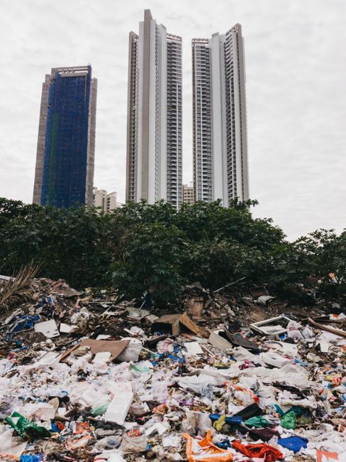 The image shows a large pile of mixed waste and rubbish, including plastic bags, cardboard boxes, and general debris, spread across an open outdoor area with some bushes and trees at the edge of the waste. In the background, there are several tall modern high-rise buildings with varying finishes; one building on the left has a blue and green façade, while the other structures are predominantly white with numerous windows. The sky above is overcast with grey clouds, and natural daylight provides diffuse lighting over the scene. The waste appears untreated and spread across a surface that might be a temporary outdoor waste collection or an unmanaged dumping site, highlighting the importance of suitable rubbish removal and waste management services such as those offered by Rubbish Collection Kennington, which specializes in private disposal and on-site clearance for urban environments near The Oval Kennington.