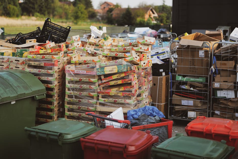 An outdoor scene showing a large pile of stacked cardboard boxes, primarily featuring empty 'fresh fruits' fruit boxes. The boxes are arranged on top of each other, with some slightly askew, and display bright red, green, and yellow branding on the packaging texture. To the right side, there are open-top wire cages filled with various cardboard and paper-based packaging, some with visible labels and printed markings. In the foreground, several large plastic bins with lids—green and red—are positioned on a paved surface, with a small shopping cart and miscellaneous loose items like a blue plastic bag and a white object nearby. Background foliage and a rural landscape with trees and houses are visible under natural daylight, creating an outdoor workspace setting suitable for private waste collection and rubbish clearance services, such as those offered by Rubbish Collection Kennington, for commercial rubbish removal or on-site clearance of recyclable materials and packaging waste.