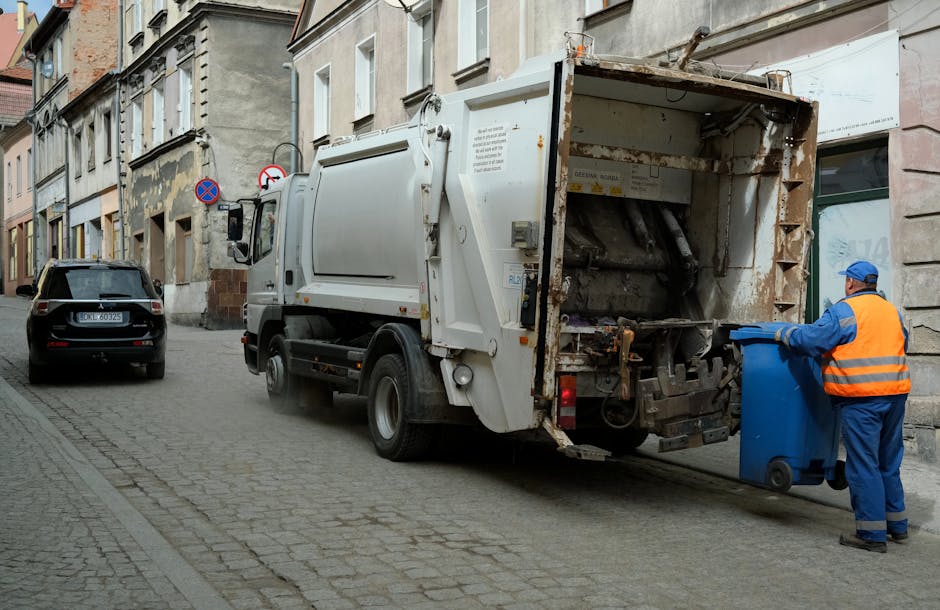 A white commercial rubbish collection truck with a rusted rear hopper is parked on a narrow cobblestone street in an urban area. The truck's rear is open, revealing internal metal components used for compacting waste. A worker dressed in a blue uniform with an orange high-visibility vest and cap is positioned on the right side, placing a blue wheelie bin into the truck's hopper, indicating an on-site waste collection process. The street is lined with older, multi-story buildings featuring stone and stucco facades, with some windows and architectural details visible above. A small, black car is parked on the left side behind the truck, and street signs indicating parking restrictions are mounted on a pole nearby. The environment suggests an operational scene of private rubbish collection as part of regular waste management servicing for urban businesses, with natural daylight and minimal shadows illuminating the scene, emphasizing the practical and functional nature of the waste disposal activity.