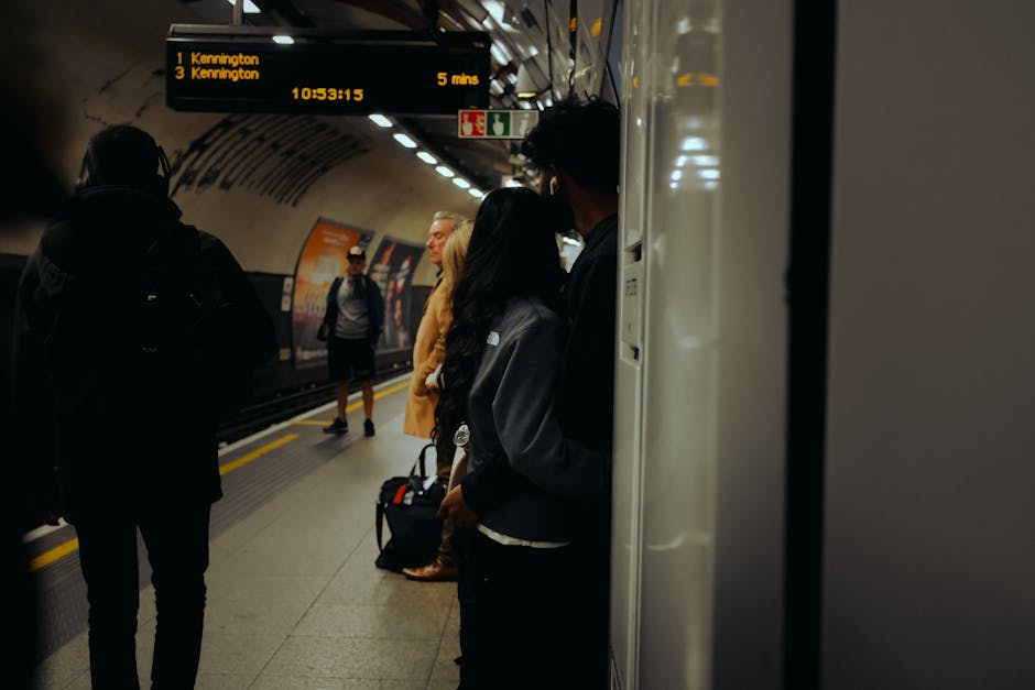 In a London underground station, several commuters are waiting on the platform, standing beside a white pillar that partially obscures some individuals. The scene shows a diverse group of people, including a woman in a black jacket holding a bag, a man wearing glasses and a hooded coat, and others dressed casually for travel. The background features a curved, tiled tunnel ceiling with embedded lighting, and the platform edge is marked with a safety yellow line. Above, a digital information display indicates train destinations and arrival times, providing essential details for travellers. The environment is illuminated by overhead lights, creating a clear, yet subdued atmosphere typical of underground transit facilities. This setting exemplifies how independent transportation hubs, like the London Tube, facilitate passenger flow and movement. For companies such as Rubbish Collection Kennington, this image highlights the importance of efficient, professional waste management in busy public spaces to maintain cleanliness and safety, even in heavily used transit environments associated with urban commuting and local services.
