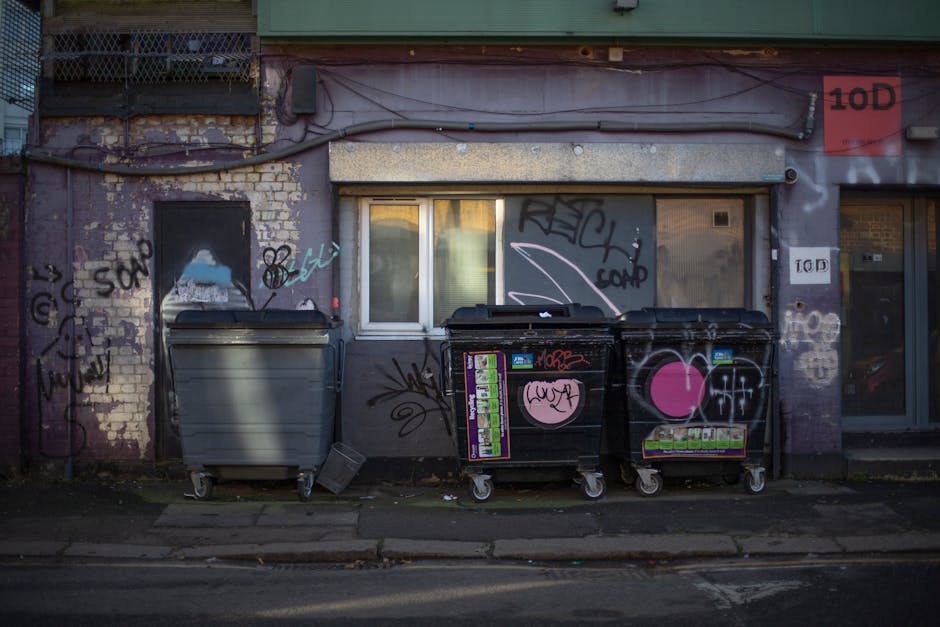 The image depicts three large wheeled rubbish containers positioned along the curbside of a building. The container on the left is dark grey with a smooth plastic surface, while the two containers on the right are black with graffiti tags and pink heart-shaped stickers on their fronts. The black containers also have various stickers and labels attached, indicating their use for waste collection. They are placed directly on the pavement in front of a building with a brick and concrete facade, which shows signs of weathering and peeling paint, especially around the window and the brickwork to the left. The window behind the containers has closed blinds, and above the window is a metal overhang or shutter mechanism. A small black object, possibly a lid or tool, lies on the ground next to the containers. The scene is lit by natural daylight, and the overall environment conveys an urban setting where independent waste disposal or collection services, such as those offered by Rubbish Collection Kennington, are used to manage local rubbish removal needs in place of council services.