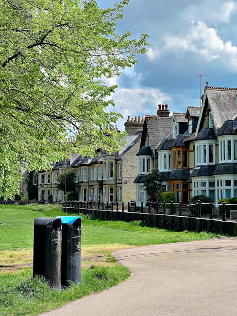 A row of traditional terraced houses with bay windows lining a residential street, featuring brick facades and slate roofs under a partly cloudy sky. In the foreground, a black outdoor rubbish bin with a blue lid is situated on a paved walkway adjacent to a grassy verge. To the left, a large leafy green tree extends its branches over the scene, providing partial shade and adding natural greenery. A low black metal fence runs parallel to the houses, separating the front gardens from the pavement. The environment appears tidy and well-maintained, reflecting a typical suburban setting where private waste collection services could be employed, as managed by companies like Rubbish Collection Kennington, for on-site clearance or alternative waste handling needs.