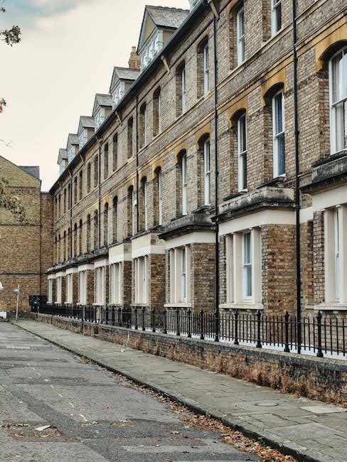 A row of traditional multi-storey residential buildings constructed from light brown and beige brick with white window frames and sills, situated along a narrow pavement. The buildings feature sash windows and small gabled roofs on the upper floors, with some windows partially open. A low black metal fence runs parallel to the sidewalk, separating the buildings from the street, which has a slightly worn, uneven surface with scattered leaves and debris. The environment suggests an urban residential area, potentially part of a private property boundary for waste collection. Natural daylight illuminates the scene, casting soft shadows on the brick facade, and the overall atmosphere is quiet and orderly, reflecting typical sightlines for property owners managing waste disposal or arranging for private rubbish removal services such as those provided by Rubbish Collection Kennington.
