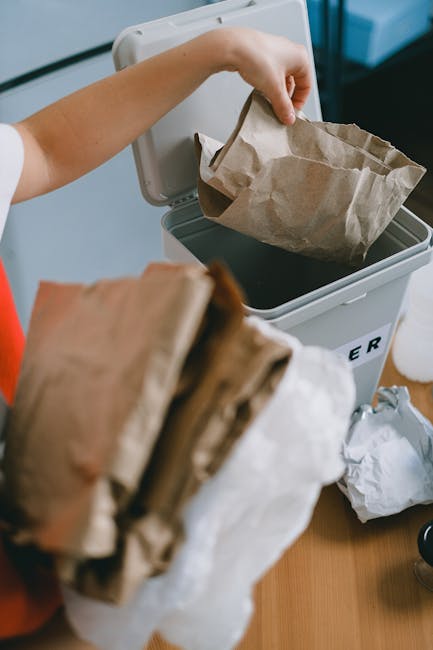 A person’s hand is seen placing a crumpled brown paper bag into a grey rubbish bin labeled 'RUBBISH'. The bin is positioned on a wooden surface, with some discarded paper and packaging next to it. The background shows a blue-toned interior environment, possibly a kitchen or office space. The paper bag appears to be used for waste disposal, reflecting typical practices of private rubbish collection and on-site clearance services provided by companies like Rubbish Collection Kennington. The scene emphasizes cleanliness and organized waste management, with a focus on recyclable or general waste materials in a controlled setting, suitable for independent rubbish removal tasks. Lighting is bright and natural, highlighting the textures of the paper bag and surrounding objects, which include crumpled waste and a partly visible white container lid. This image illustrates the process of sorting and disposing of waste efficiently in a domestic or small commercial environment, aligning with professional rubbish removal services often utilized in urban areas for alternative waste handling.