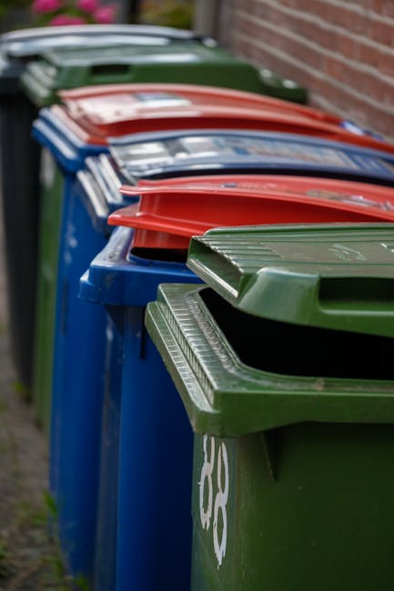 A row of large, rectangular wheelie bins lined up along a pavement near a brick wall, with green, red, and blue lids facing outward. The bins are made of durable plastic with textured surfaces and prominent lids designed for rubbish disposal. The green and red-lidded bins are positioned in the foreground, with the blue-lidded bin adjacent. The background shows a slight blur of grassy area and some foliage, indicating an outdoor environment. The scene appears to represent managed waste collection, highlighting the range of container colours used for different waste types, which could relate to private rubbish collection services. The lighting is natural and even, emphasizing the clean, organized presentation of waste containers, which are associated with rubbish management providers such as Rubbish Collection Kennington, serving local communities with alternative waste handling solutions.