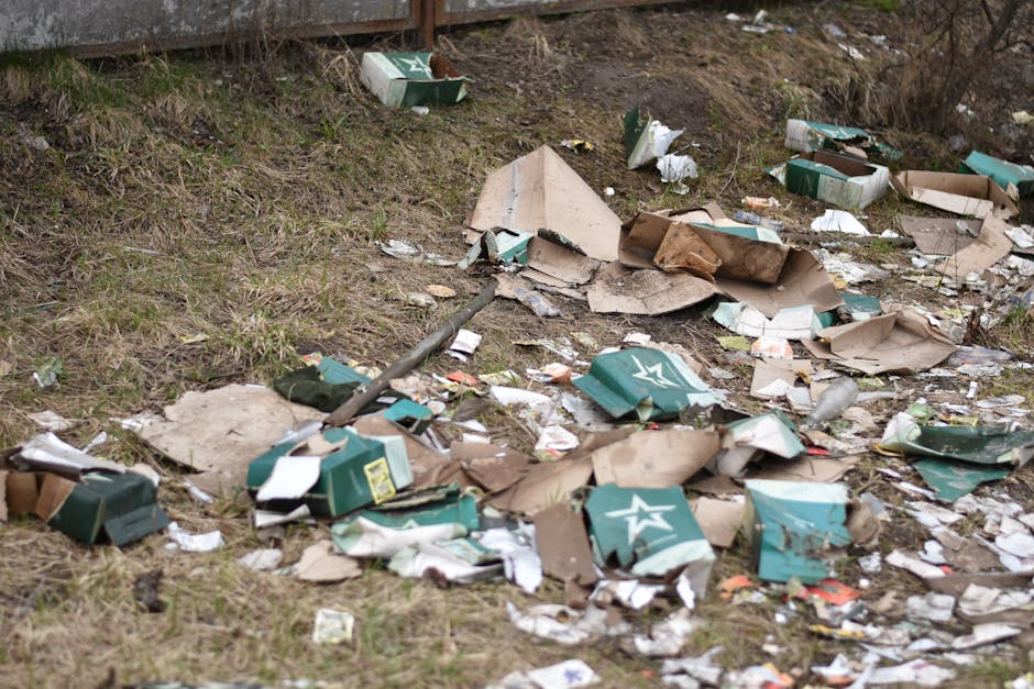 Photograph of an outdoor area showing scattered discarded cardboard boxes, some flattened and torn, with visible green and white branding, mixed with crumpled paper and plastic packaging, all spread across a patchy grassy surface next to a worn concrete structure at the back. The boxes and debris are haphazardly strewn across the ground, with some partially resting on the grass and others overlapping. The scene is illuminated by natural daylight, highlighting the textures of the paper, cardboard, and grass, creating a cluttered and neglected appearance characteristic of illegal fly tipping on a roadside or private land. The presence of rubbish and scattered waste materials demonstrates an example of improper waste disposal, emphasizing the importance of proper rubbish management and alternative waste handling services, such as those offered by Rubbish Collection Kennington, to prevent illegal dumping in residential or natural areas.