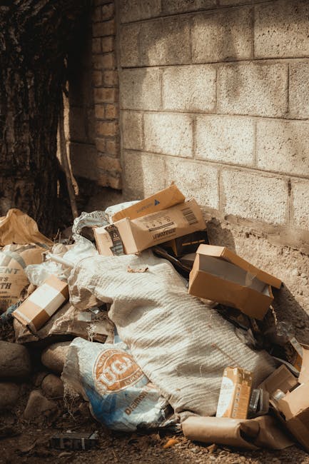 A pile of discarded cardboard boxes, some with open flaps, and large white and beige sacks of rubbish located at the base of a stone and brick wall, adjacent to a tree trunk. The boxes appear to be used for packaging and are partially crushed, with visible tape and printed labels. The sacks are textured, made from woven plastic or cloth, and are filled with waste materials, bulging at the seams. The scene is outdoors, possibly in a private or communal area, with natural lighting highlighting the materials' surfaces. The background features a brick wall to the right and a rough, stony ground beneath the rubbish. This setup resembles a typical example of unmanaged waste accumulation, which could be addressed through private rubbish collection services, such as those offered by Rubbish Collection Kennington, for proper disposal or clearance, avoiding fly tipping or illegal dumping in residential or urban environments.