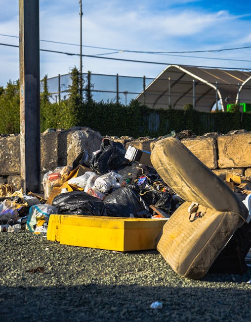 Photograph of an outdoor area showing scattered discarded cardboard boxes, some flattened and torn, with visible green and white branding, mixed with crumpled paper and plastic packaging, all spread across a patchy grassy surface next to a worn concrete structure at the back. The boxes and debris are haphazardly strewn across the ground, with some partially resting on the grass and others overlapping. The scene is illuminated by natural daylight, highlighting the textures of the paper, cardboard, and grass, creating a cluttered and neglected appearance characteristic of illegal fly tipping on a roadside or private land. The presence of rubbish and scattered waste materials demonstrates an example of improper waste disposal, emphasizing the importance of proper rubbish management and alternative waste handling services, such as those offered by Rubbish Collection Kennington, to prevent illegal dumping in residential or natural areas.